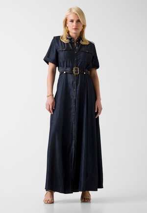 Kobieta w ciemnoniebieskiej długiej sukience z dżinsu z paskiem, krótkimi rękawami, złotymi kolczykami, bransoletką i beżowymi sandałami na obcasie, stojąca na białym tle.