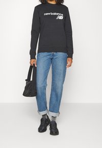 Svart sweatshirt med vit "new balance" logotyp, blå raka jeans, svarta sneakers och svart väska i texturerad tyg.