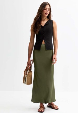 Haut sans manches noir à côtes avec un devant à boutons, associé à une jupe maxi olive ajustée. Le mannequin tient un sac en osier.