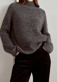 Pull en tricot gris à côtes avec un col haut, featuring des manches amples, porté avec un pantalon en velours côtelé noir. Texture douce avec un motif de tricot très marqué.