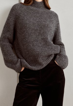 Pull en tricot gris à côtes avec un col haut, featuring des manches amples, porté avec un pantalon en velours côtelé noir. Texture douce avec un motif de tricot très marqué.