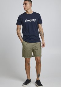 Temno modra bombažna majica s besedilom "simplify." v beli barvi, v kombinaciji s khaki kratkimi hlačami z zavitimi robovi, obuta s črnimi teniskami.