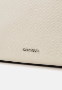 Surface en tissu beige texturé avec un petit logo Calvin Klein argenté centré près du bord inférieur.