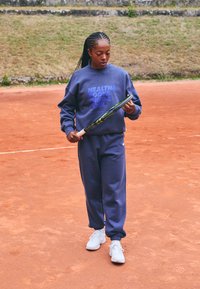 Sweatshirt bleu marine et pantalon de survêtement assorti en mélange de coton, avec texte imprimé. Chaussures de sport blanches, tenant une raquette de tennis sur un court de terre battue.