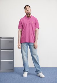 Camisa polo rosa de manga corta con un pequeño logo, combinada con jeans de color azul claro y zapatillas blancas. Fondo neutro y archivo gris.