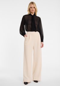 Zwarte sheer blouse met een gestrikt detail bij de hals, gecombineerd met hoge taille, crèmekleurige wijde broekspijpen. Beide kledingstukken hebben een gladde textuur.