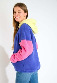 Felpa in pile con corpo blu, maniche rosa e cappuccio giallo. Presenta una vestibilità comoda e tasche a marsupio sul davanti.
