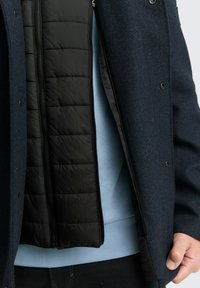 Sacou herringbone de culoare bleumarin, cu căptușeală neagră quilted, având închidere prin fermoar și nasturi tip snaph, purtat peste un pulover de culoare bleu deschis.