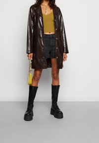 Hnědý lesklý kabát z umělé kůže, černé džíny šortky, zelený pletený crop top a robustní černé šněrovací boty; drží malou žlutou tašku.