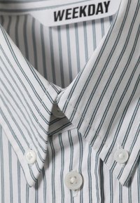 Camisa branca de vestir com finas riscas verticais pretas, colarinho com botões e uma etiqueta com a inscrição "WEEKDAY" dentro do colarinho.