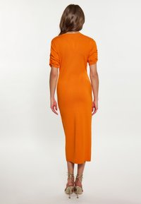 Rochie midi tricotată portocalie, cu mâneci scurte răsucite, siluetă ajustată și tiv drept. Însoțită de pantofi cu toc înalt bej, cu barete.