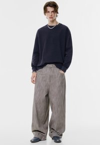 Marinefarbener Langarm-Sweatshirt kombiniert mit weitem, hellgrau gestreiften Hosen. Das Model trägt eine silberne Kette und Sneakers. Schlicht, lockere Passform.