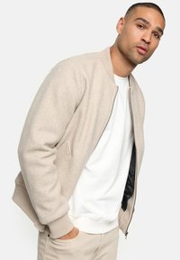 Beige bomber jacket with a soft texture, ribbed cuffs, and a zip closure. Worn over a white t-shirt, features side pockets and a black lining.