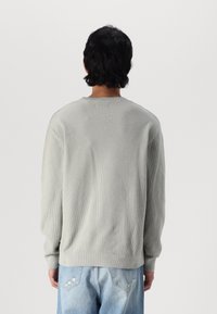 Persoon met kort zwart haar, gekleed in een lichtgrijze gebreide trui en lichtblauwe spijkerbroek, staande met de rug naar de camera.