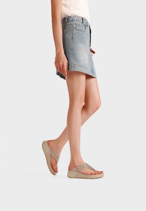 Persona che indossa una minigonna in denim azzurro chiaro e sandali beige con zeppa e motivo decorativo traforato, in piedi su sfondo bianco.