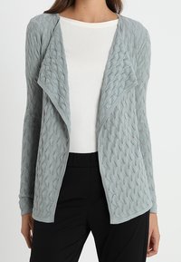 Ljust blå stickad cardigan med öppen framsidan, med ett strukturerat diamantmönster och långa ärmar, lager på lager över en vit topp.