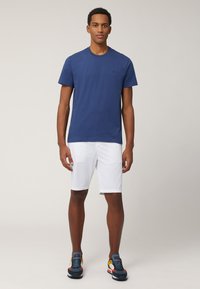 T-shirt in cotone blu navy con scollo rotondo, abbinato a pantaloni corti bianchi. Il modello indossa scarpe da ginnastica nere e blu con dettagli colorati.