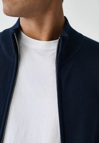 Granatowy kardigan na zamek z prążkowanym kołnierzykiem, założony na biały t-shirt. Posiada srebrny zamek, gładką fakturę oraz dopasowany krój.