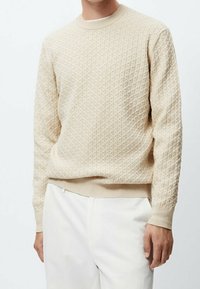 Beige gemusterter Pullover mit einem strukturierten Rautendesign, geripptem Kragen und Bündchen, kombiniert mit weißen Hosen.