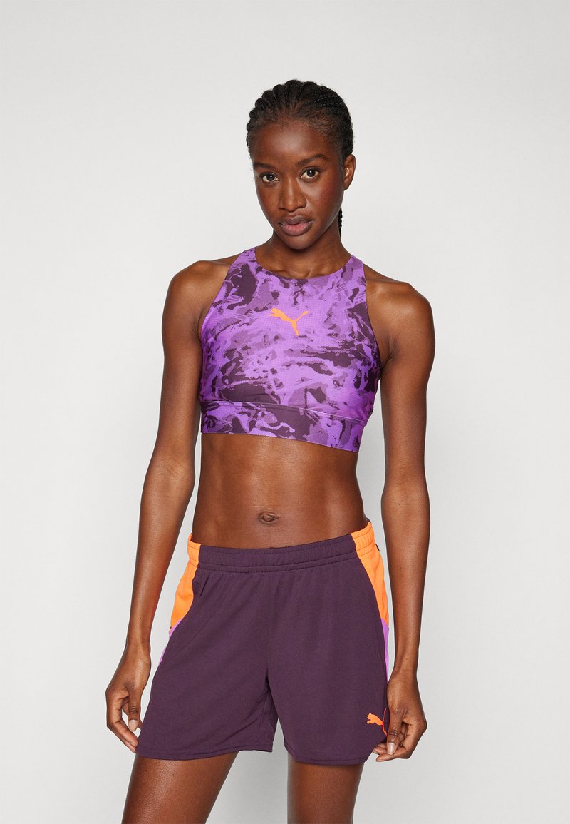 Violetti urheilu crop top, jossa on kuvio ja oranssit korostukset, yhdistettynä tummansinisiin shortsit, joissa on oranssit sivupaneelit ja logo.