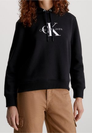 Sudadera de algodón negra con un diseño de logo en blanco en la parte frontal, con capucha con cordón y puños y dobladillo de canalé. Combinada con pantalones cargo beige.