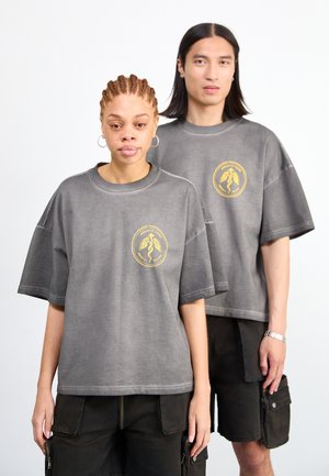 Zwei Models tragen übergroße, graue Baumwoll-T-Shirts mit einem goldenen Grafiklogo. Sie sind mit dunklen Cargo-Shorts gestylt.