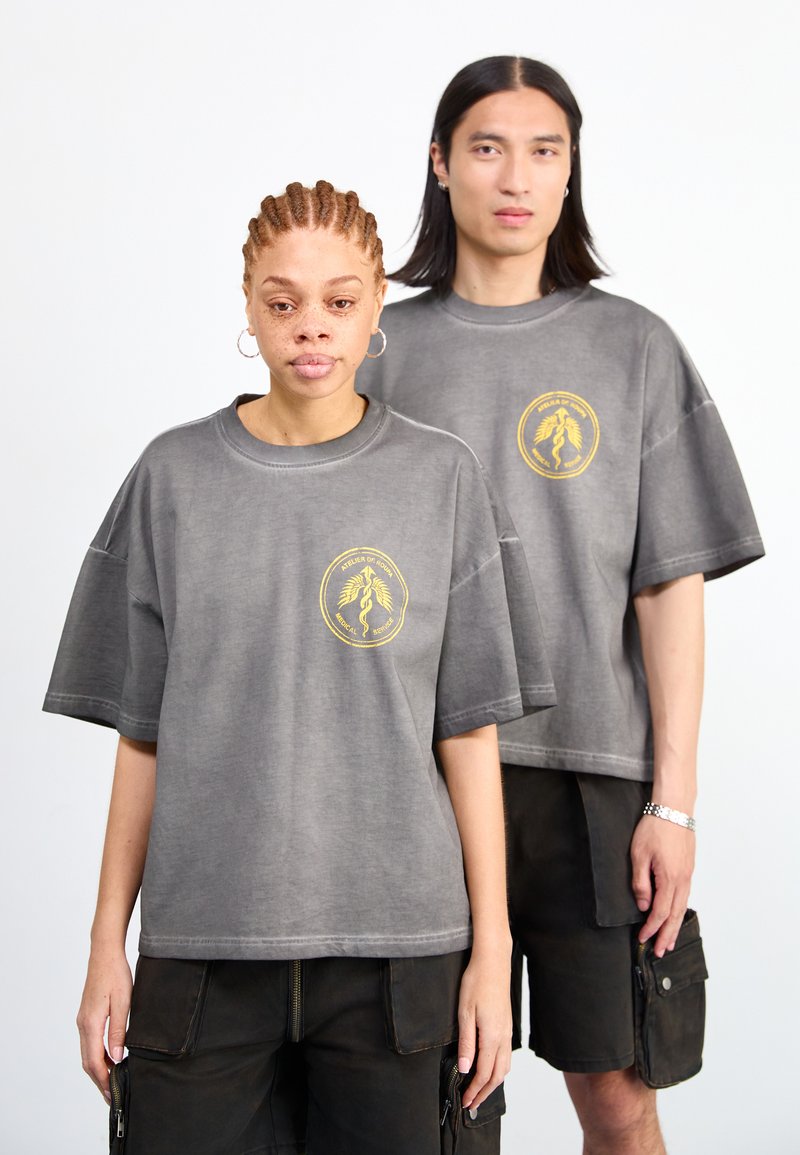 Dos modelos llevan camisetas de algodón gris de gran tamaño con un logo gráfico dorado. Están combinadas con pantalones cortos oscuros de cargo.