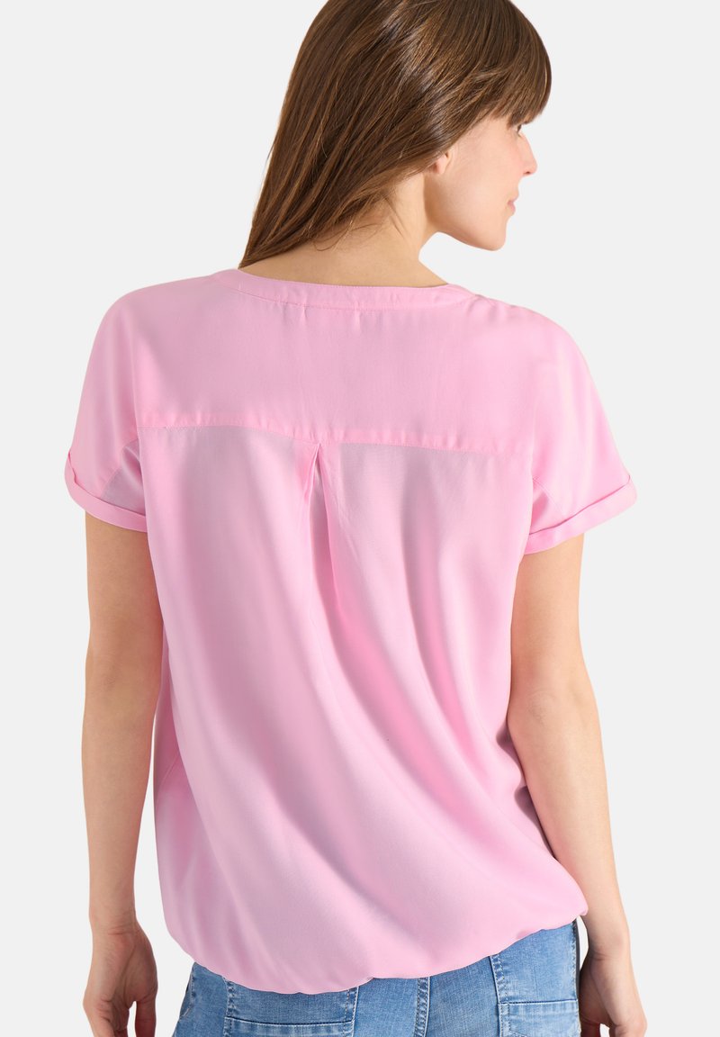 Femme aux longs cheveux bruns portant un chemisier rose à manches courtes et amples ainsi qu'un jean bleu, vue de dos sur un fond uni.