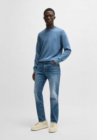 Helles blaues Sweatshirt mit Rundhalsausschnitt und kleinem Logo, kombiniert mit mittelblauen Jeans und beigen Sneakers. Lässiges Outfit.