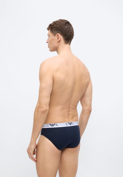 Noormees, keda nähakse tagantpoolt, kannab tumesiniseid aluspesu valge kaubamärgiga vöökohaga ja seisab ühevärvilise helehalli tausta ees.