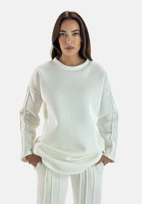 Biała bluza z okrągłym dekoltem i ściągaczami, o luźnym kroju. W połączeniu z dopasowanymi białymi spódnicami plisowanymi. Miękka, teksturowana tkanina.