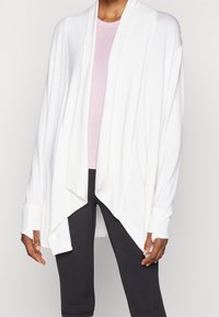 Cardigan bianco aperto davanti realizzato in tessuto morbido, con una vestibilità comoda, maniche lunghe e orlo asimmetrico. Indossato sopra un top di un rosa chiaro.