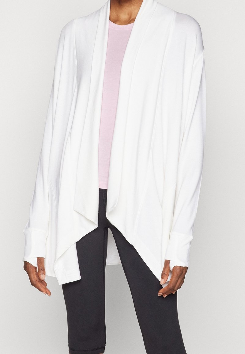 Cardigan bianco aperto davanti realizzato in tessuto morbido, con una vestibilità comoda, maniche lunghe e orlo asimmetrico. Indossato sopra un top di un rosa chiaro.