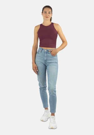 Frau, die in einem ärmellosen kastanienbraunen Crop-Top, hochgeschnittenen hellblauen Jeans und weißen Sneakers steht, mit einer Hand in ihrer Tasche.