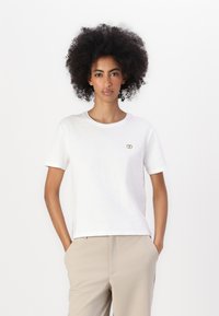T-shirt bianca a maniche corte in cotone con scollatura rotonda e un piccolo logo dorato sul petto. Indossata con pantaloni beige.