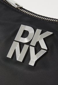 Strieborné kovové logo DKNY na čiernej textúrovanej látke pri striebornom zipsu.