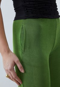 Kvinnas hand med guldring nära fickan på gröna byxor, iklädd en svart rynkad topp med prickar, mot en ljus enfärgad bakgrund.