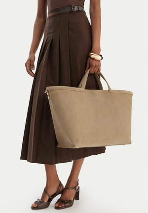 Frau trägt einen dunkelbraunen, plissierten Midirock und Sandalen, hält eine große beigefarbene Tragetasche mit kurzen Griffen und trägt ein goldenes Armband am Handgelenk.