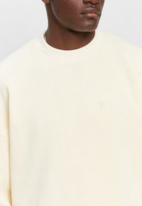 Crème kleurige sweatshirt met een gewatteerd wafelbreipatroon, ronde ronde halslijn en een klein, subtiel logo op de borst.