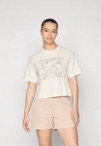 Cremfarbener, verkürzter T-Shirt mit Grafikdruck, Rundhalsausschnitt und kurzen Ärmeln, kombiniert mit beigen, gerippten Shorts, die eine glatte Textur aufweisen.