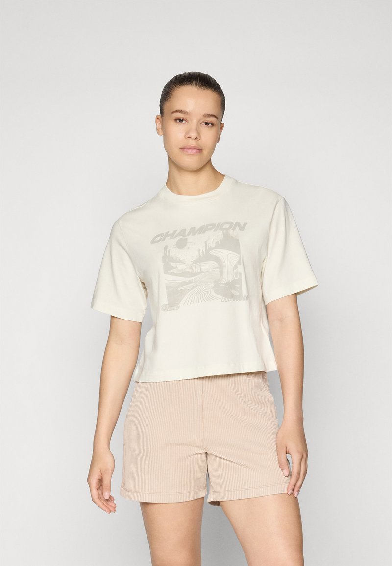 Cremfarbener, verkürzter T-Shirt mit Grafikdruck, Rundhalsausschnitt und kurzen Ärmeln, kombiniert mit beigen, gerippten Shorts, die eine glatte Textur aufweisen.