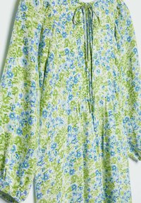 Langärmliges Kleid mit grünem und blauem Blumenmuster, geraffter Taille und einer Bindung am Ausschnitt auf hellem Stoffhintergrund.