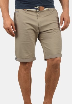 Mand iført beige rullede shorts til knæet med et vævet bælte og en marineblå skjorte, stående mod en ensfarvet baggrund.