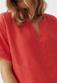 Žena s blond vlasy po ramena, oblékající červený lněný top s krátkým rukávem a výstřihem do V na bílém pozadí.