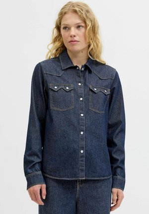 Camicia in denim blu scuro con maniche lunghe, chiusura a bottoni a pressione, colletto a punta e tasche sul petto doppie. Presenta accenti di cuciture a vista.