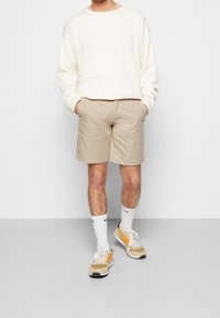 Beige shorts en een crèmekleurige rib gebreide trui, aangevuld met witte sokken en beige sneakers met oranje accenten. Eenvoudige, casual outfit.