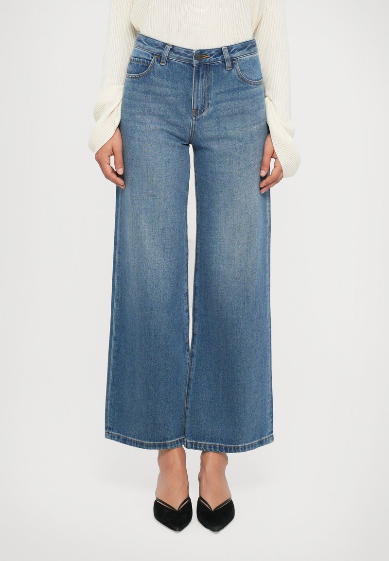 Mulher vestindo jeans azuis de cintura alta e perna larga, suéter creme canelado de mangas longas e sapatos pretos de bico fino, em pé contra um fundo branco.