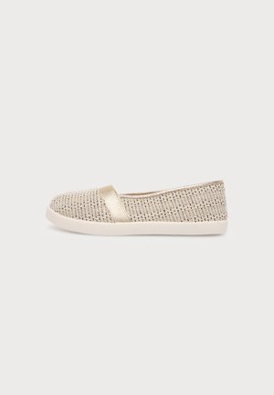 Beige vævet slip-on flad sko med hvid sål og en metallisk guldrem over vristen, vist i profil på en ensfarvet baggrund.