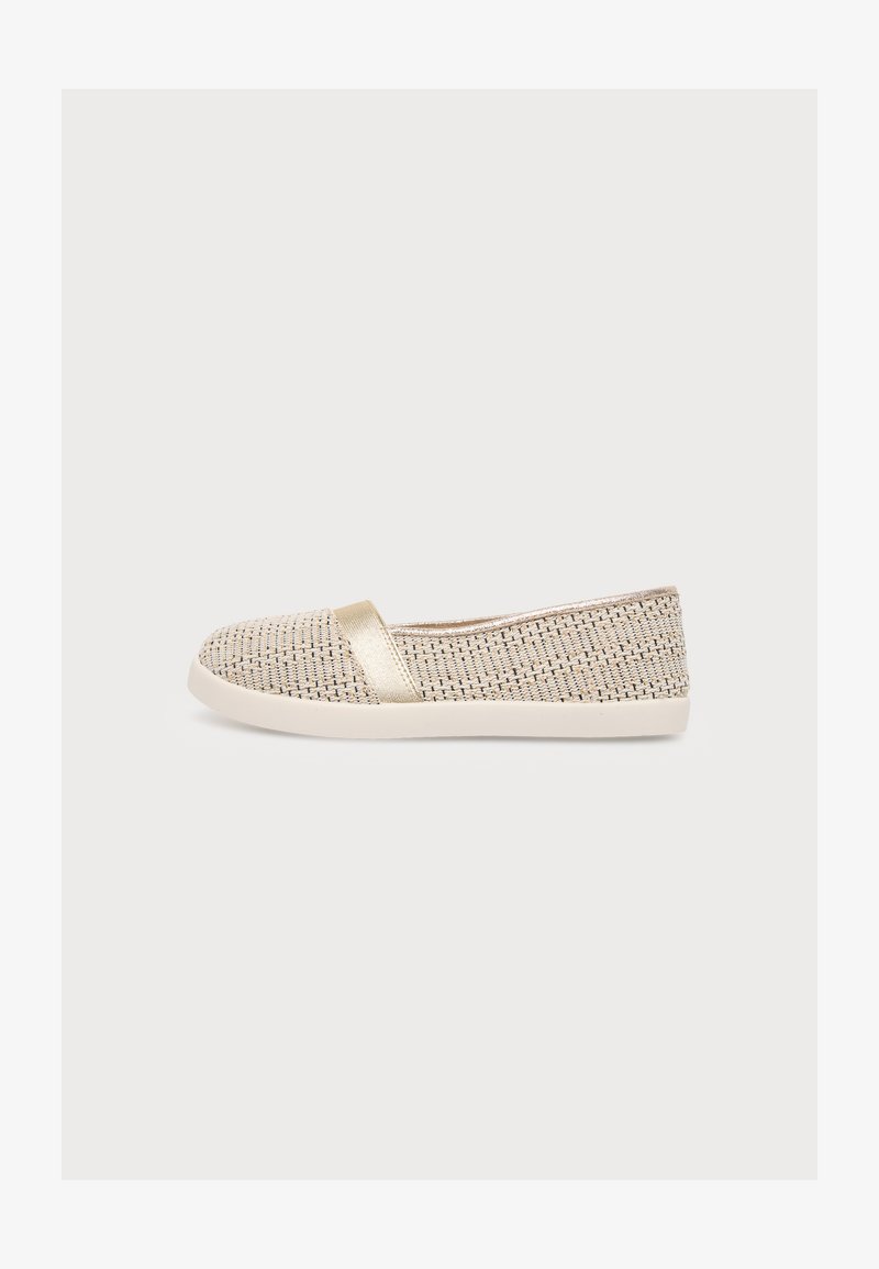 Μπεζ υφασμάτινο επίπεδο slip-on παπούτσι με λευκή σόλα και μεταλλική χρυσή λωρίδα στο επάνω μέρος, εμφανίζεται σε προφίλ σε απλό φόντο.