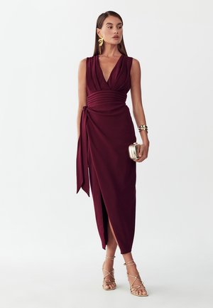 Mujer con vestido sin mangas color burdeos oscuro cruzado con abertura, tacones dorados de tiras, grandes pendientes de hoja dorados, pulsera dorada y sosteniendo un pequeño bolso de mano dorado.